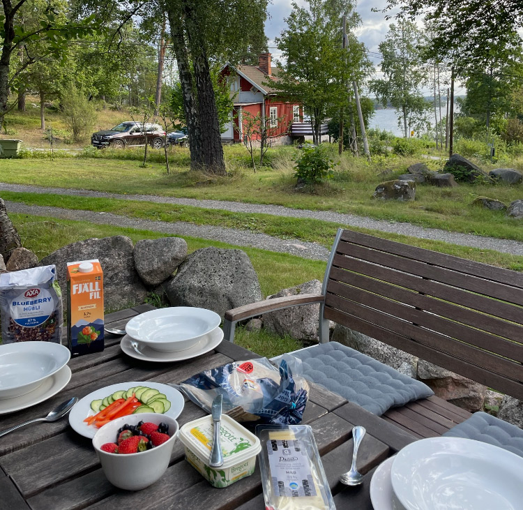 Outdoor breakfast setting with red farmhouse in background, fresh fruits, bread, cheese, yogurt, and muesli.